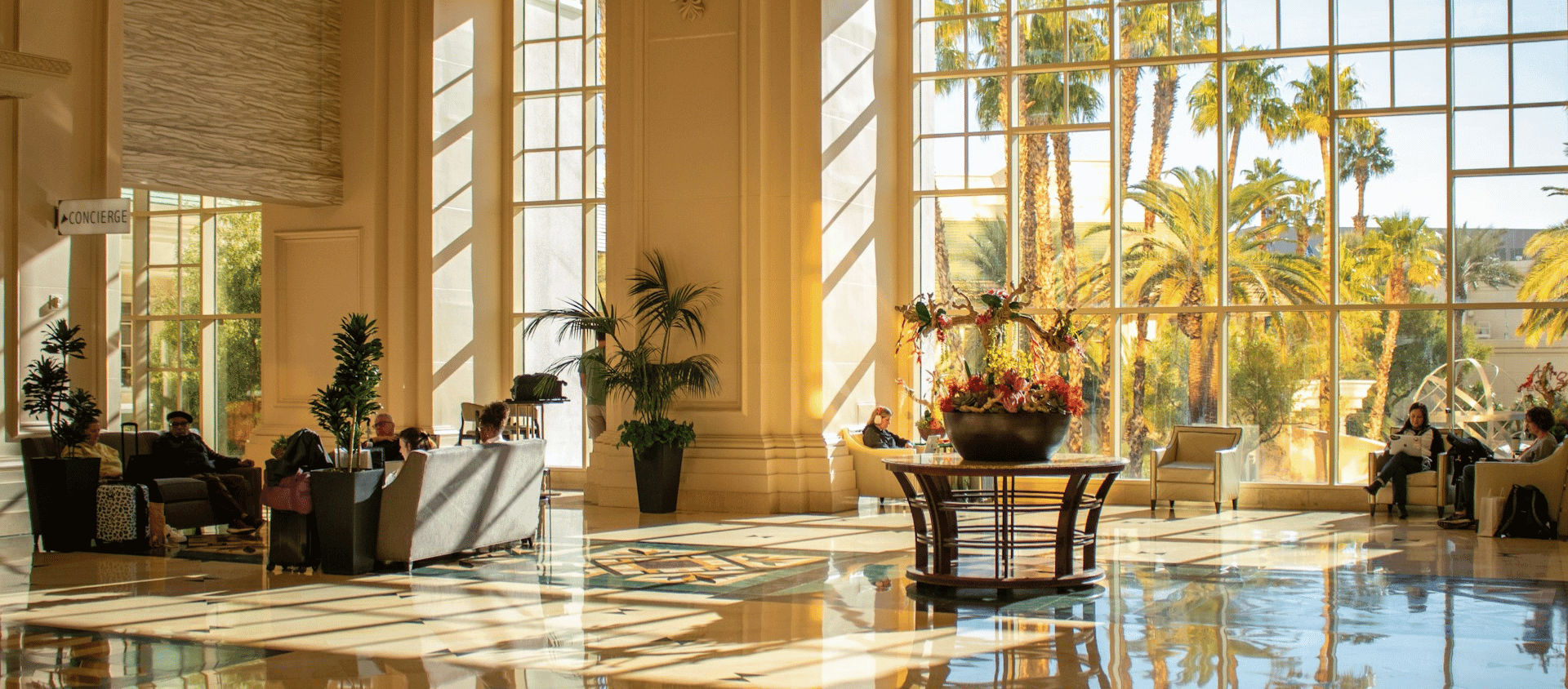 Sunlit grand hotel lobby with towering windows and lush palm trees.