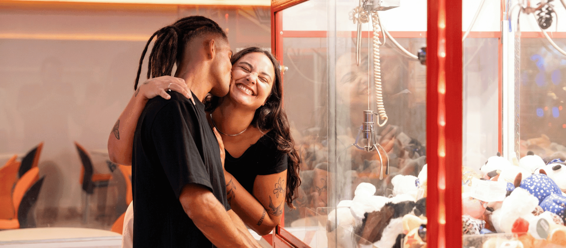 A couple hugging and smiling next to a claw machine filled with plush toys.