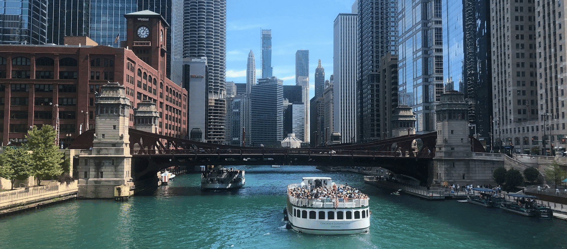 Chicago River with a sightseeing boat passing under a bridge surrounded by skyscrapers.