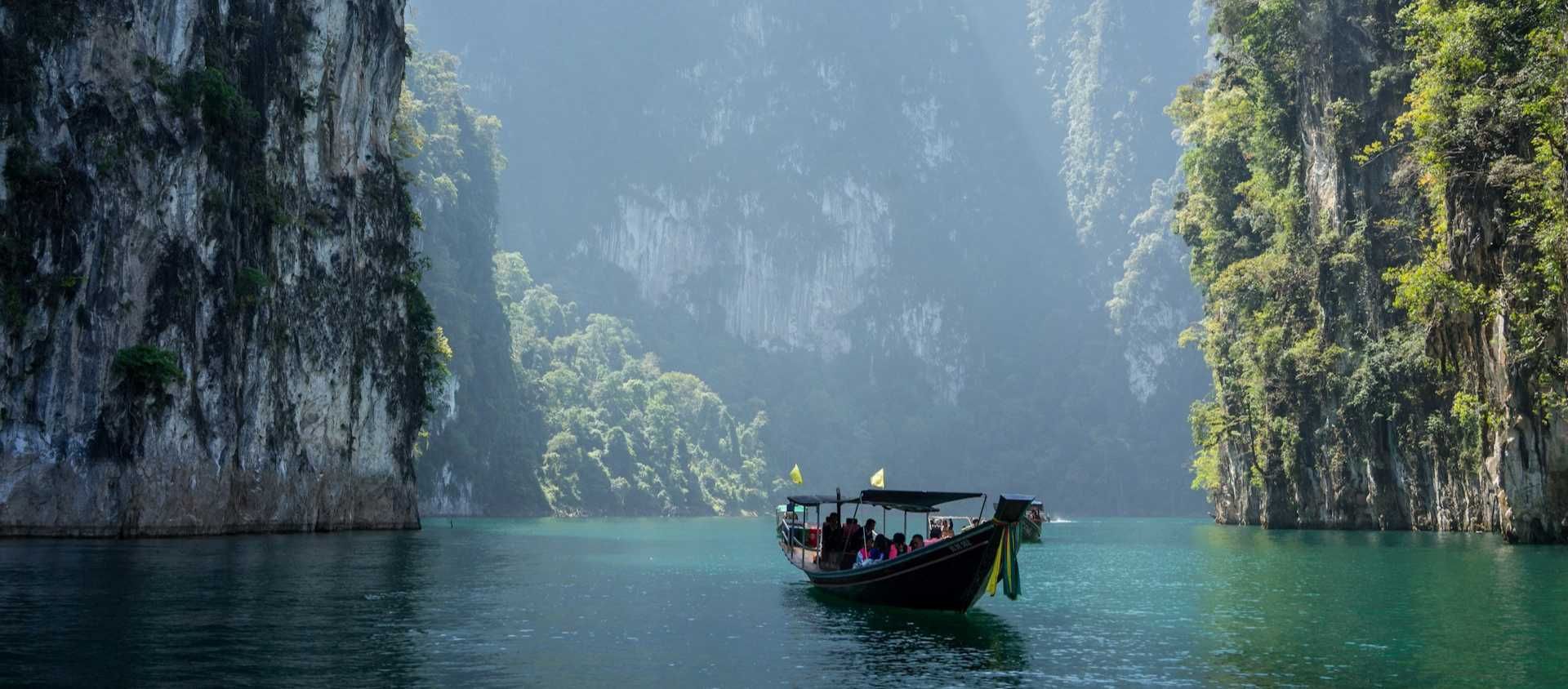 Longtail boat passing towering limestone cliffs on emerald water in Thailand.