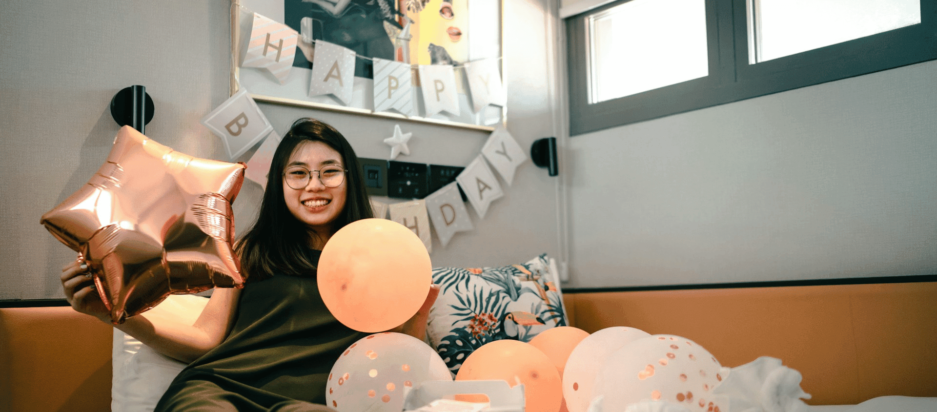 A woman holding balloons in a room decorated with a birthday banner.