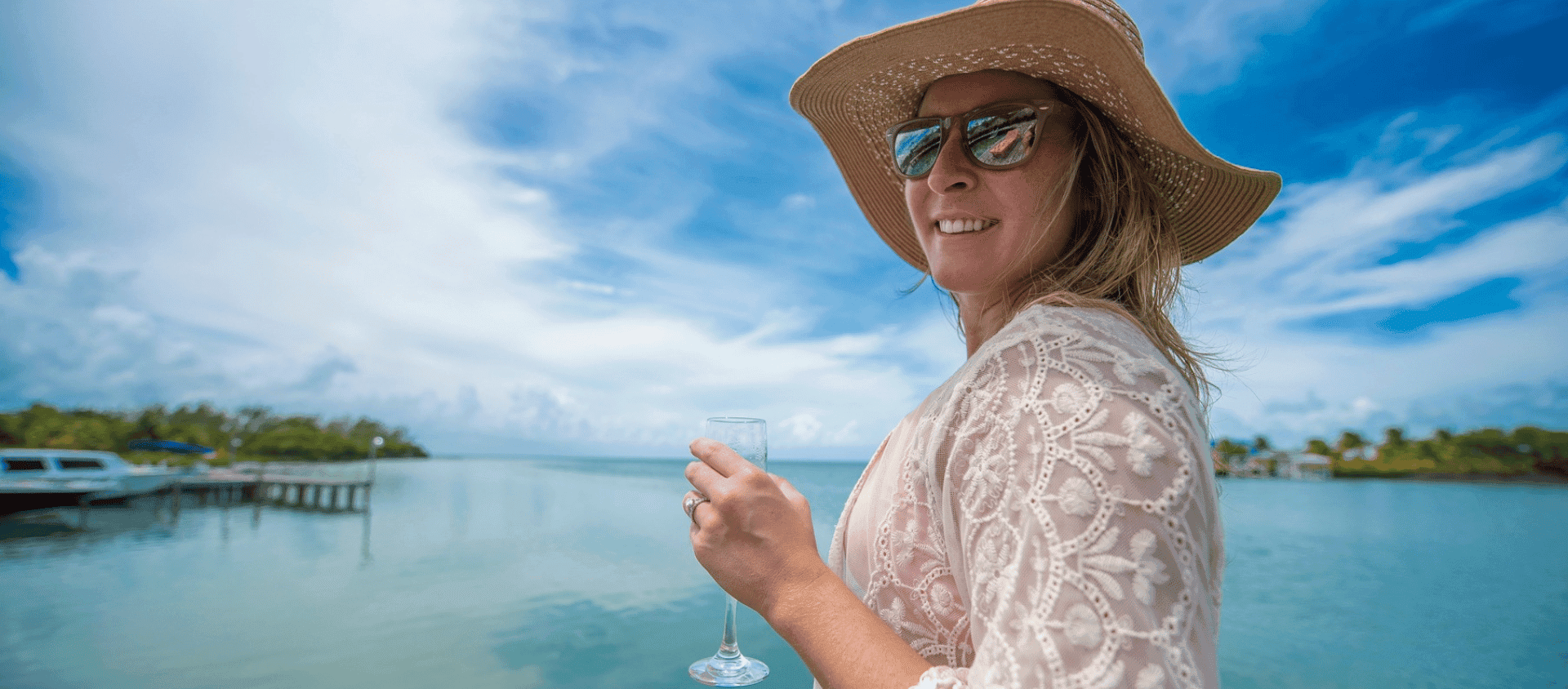 A woman wearing a sun hat and sunglasses holds a drink while standing by calm ocean water under a blue sky.