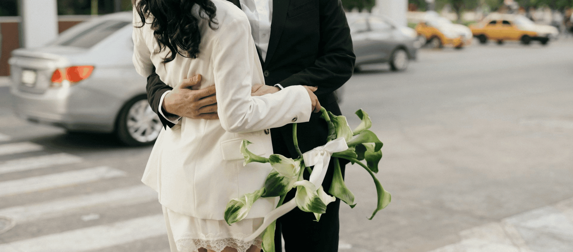 Couple in wedding attire embracing on a city street, holding a bouquet of white flowers.