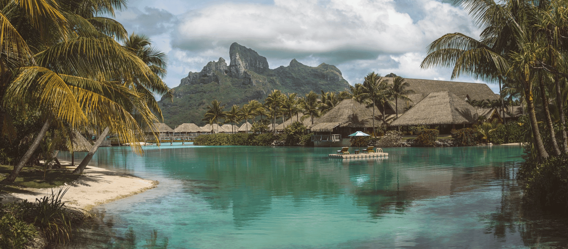 Palm lined lagoon in Bora Bora with turquoise water, traditional thatched bungalows, and a mountain under cloudy skies.