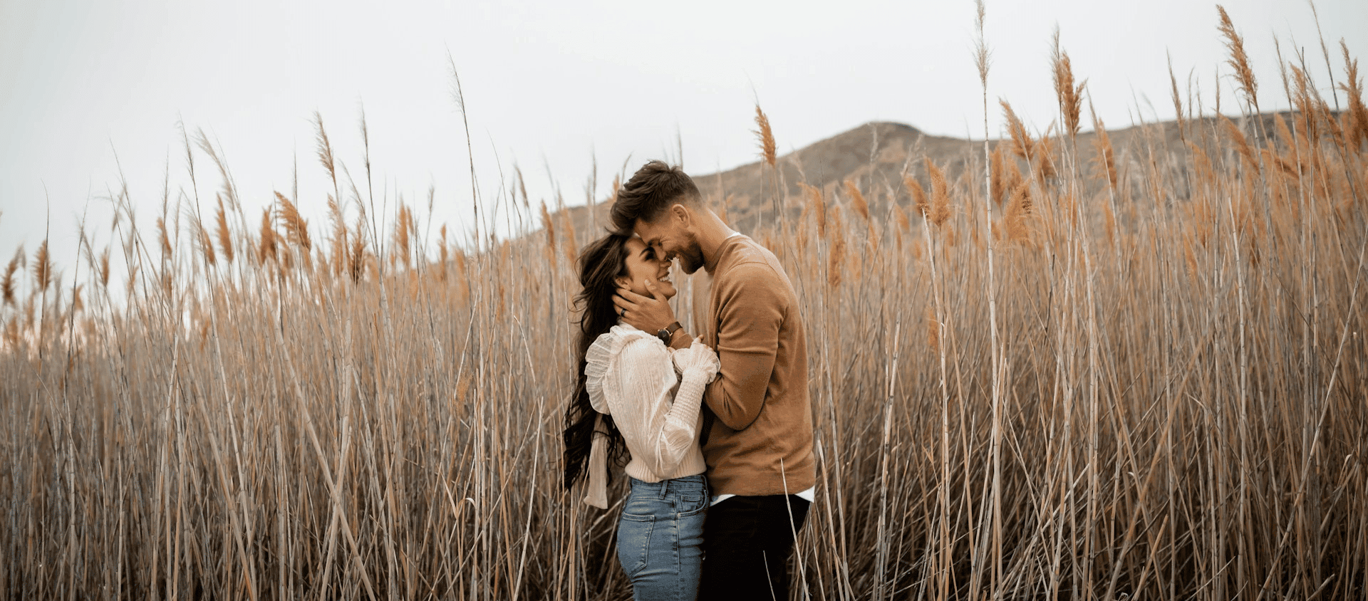 Couple embracing in a field of tall grass with mountains in the background.