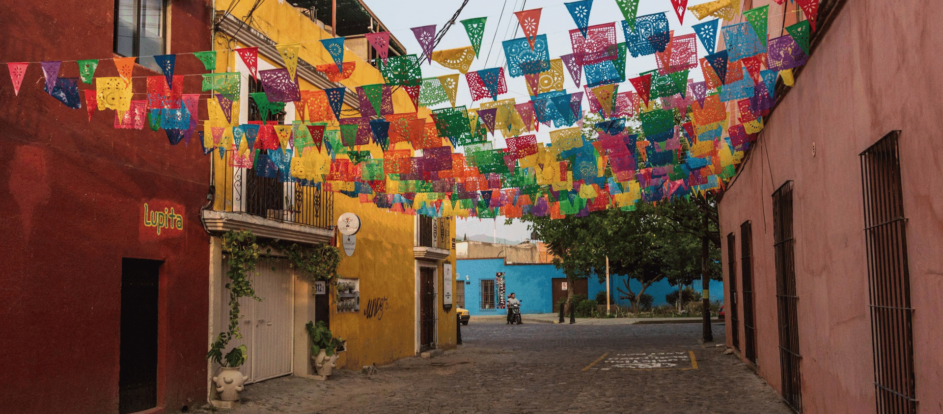 Colorful papel picado banners hanging above a cobblestone street lined with vibrant buildings