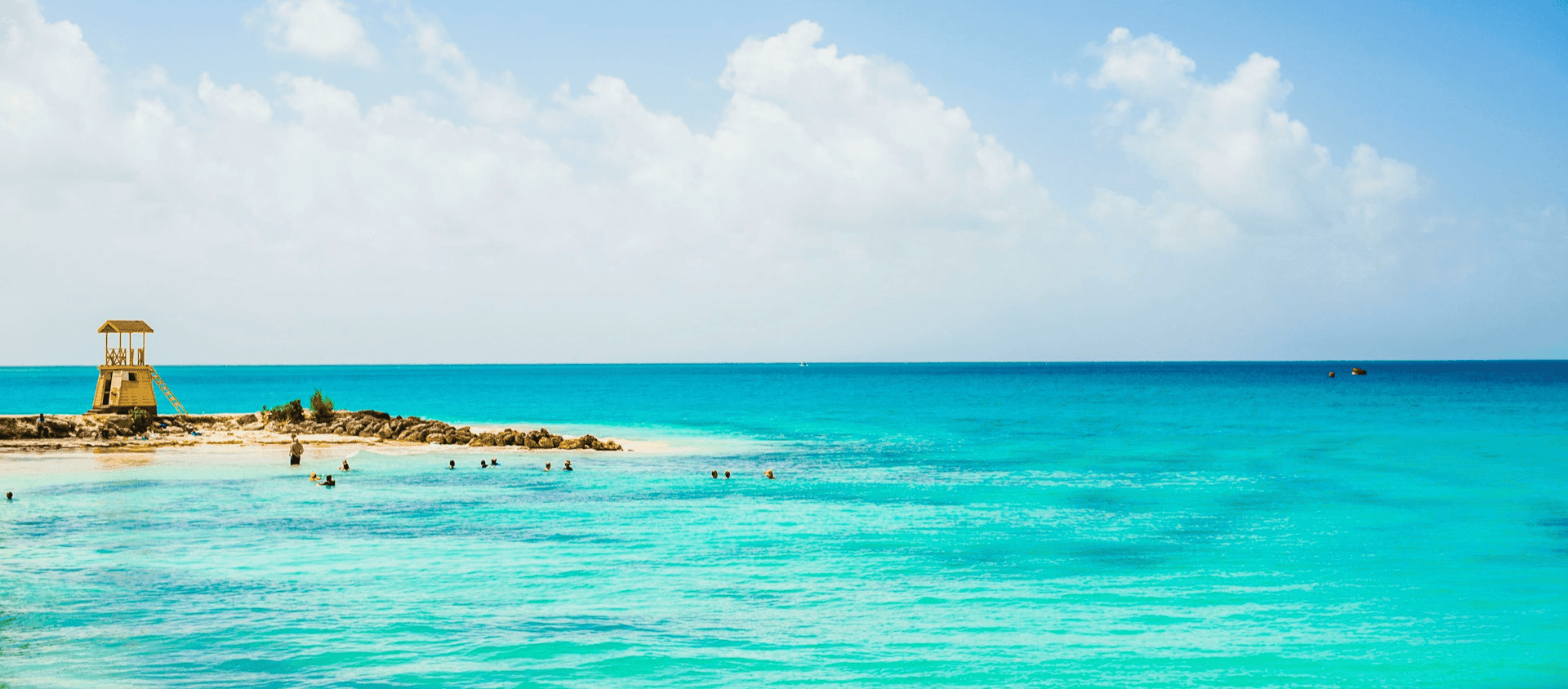Brilliant turquoise water meeting pale sand on the South Coast of Barbados, with a small lifeguard hut on a rocky outcrop.