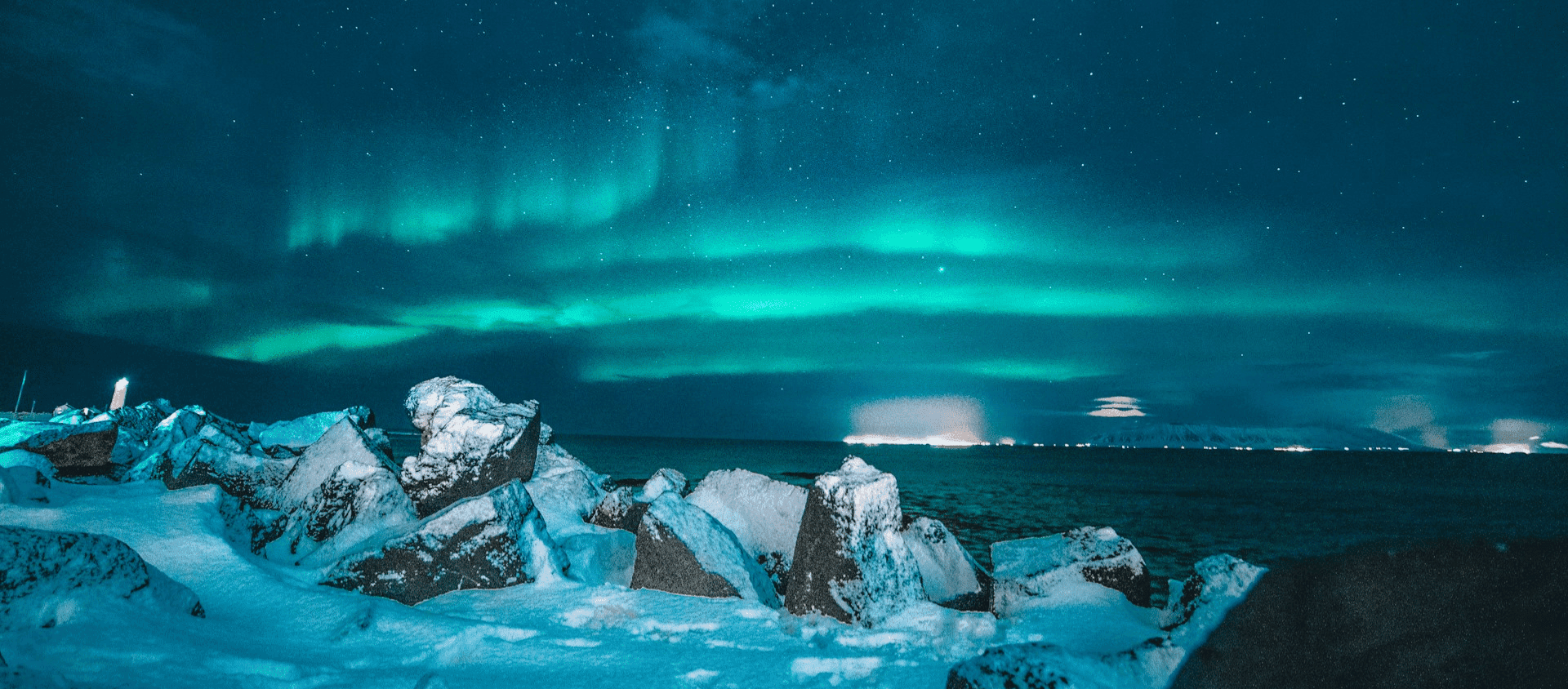 Snow-covered rocks by the sea under bright green northern lights.
