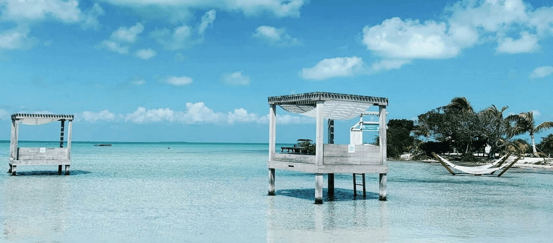 An overwater cabana on stilts above calm turquoise Caribbean water near a caye in Belize, with a hammock strung between palms.