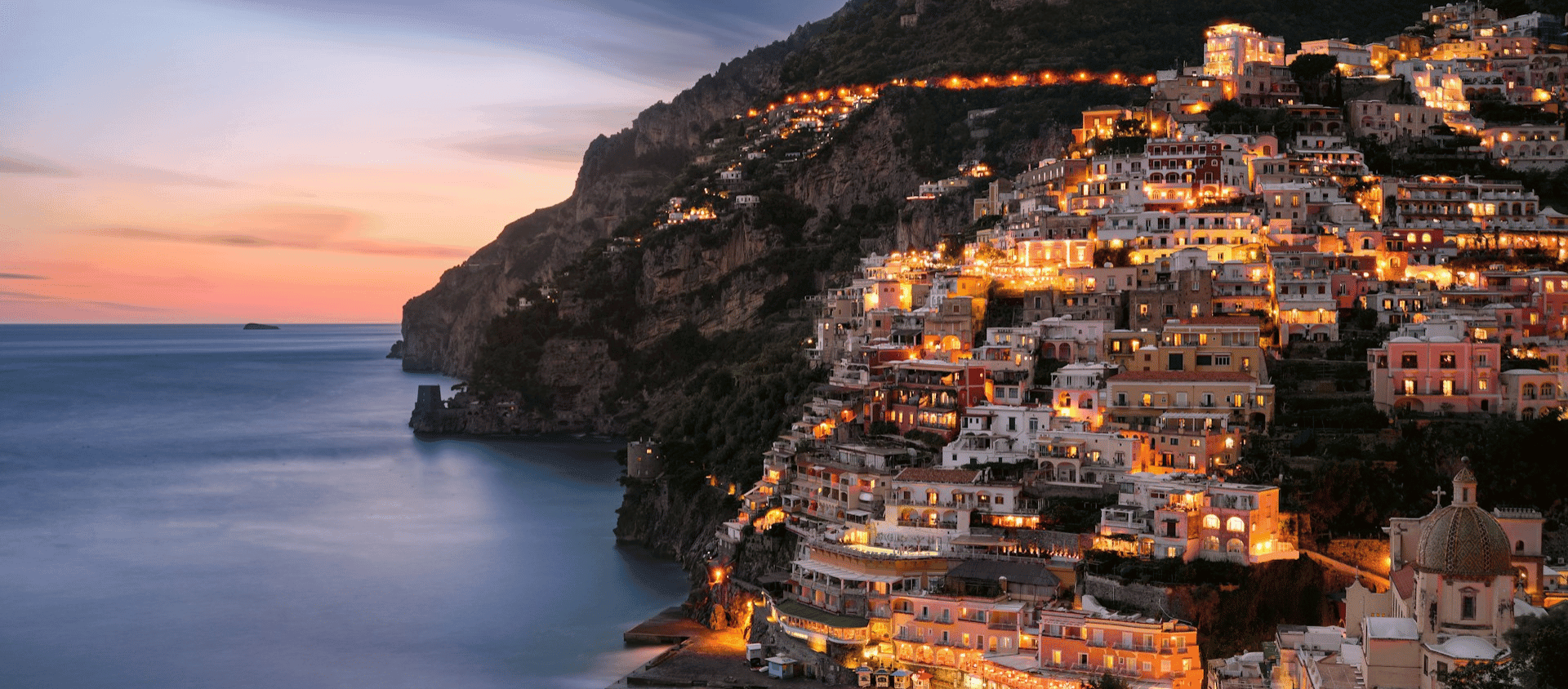 Positano’s cliffside homes glowing at sunset above the calm Mediterranean shoreline.