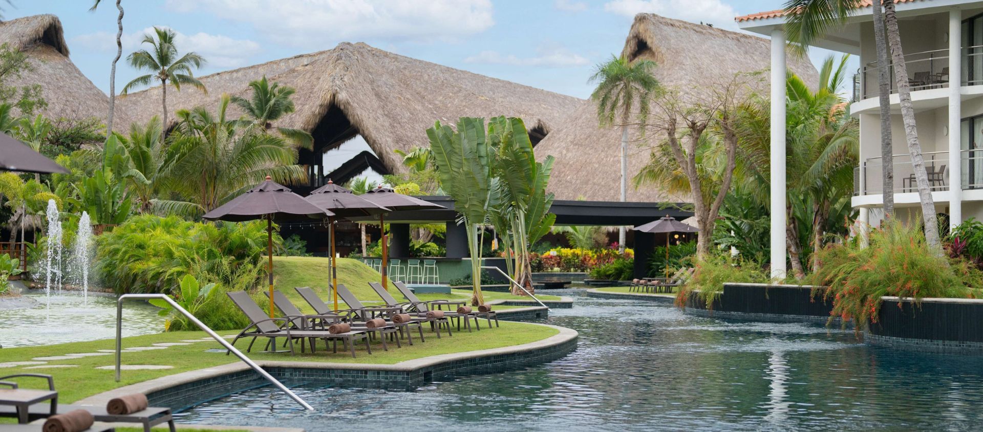 Resort pool surrounded by tropical landscaping, lounge chairs, and thatched-roof cabanas.