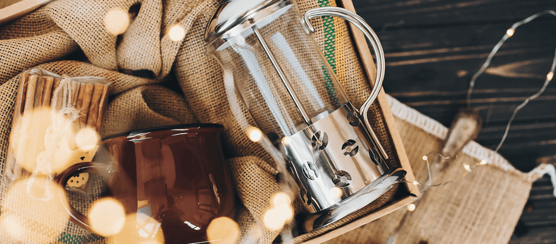 Coffee press, mug, and cookies arranged on burlap fabric with warm lights.