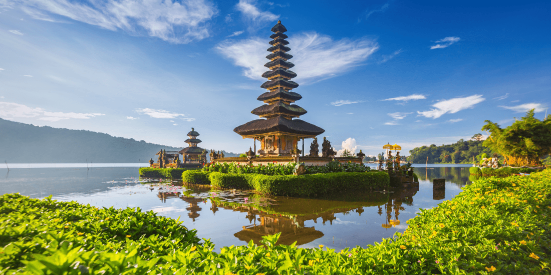 Multi-tiered lakeside temple reflected in calm water under a blue sky.