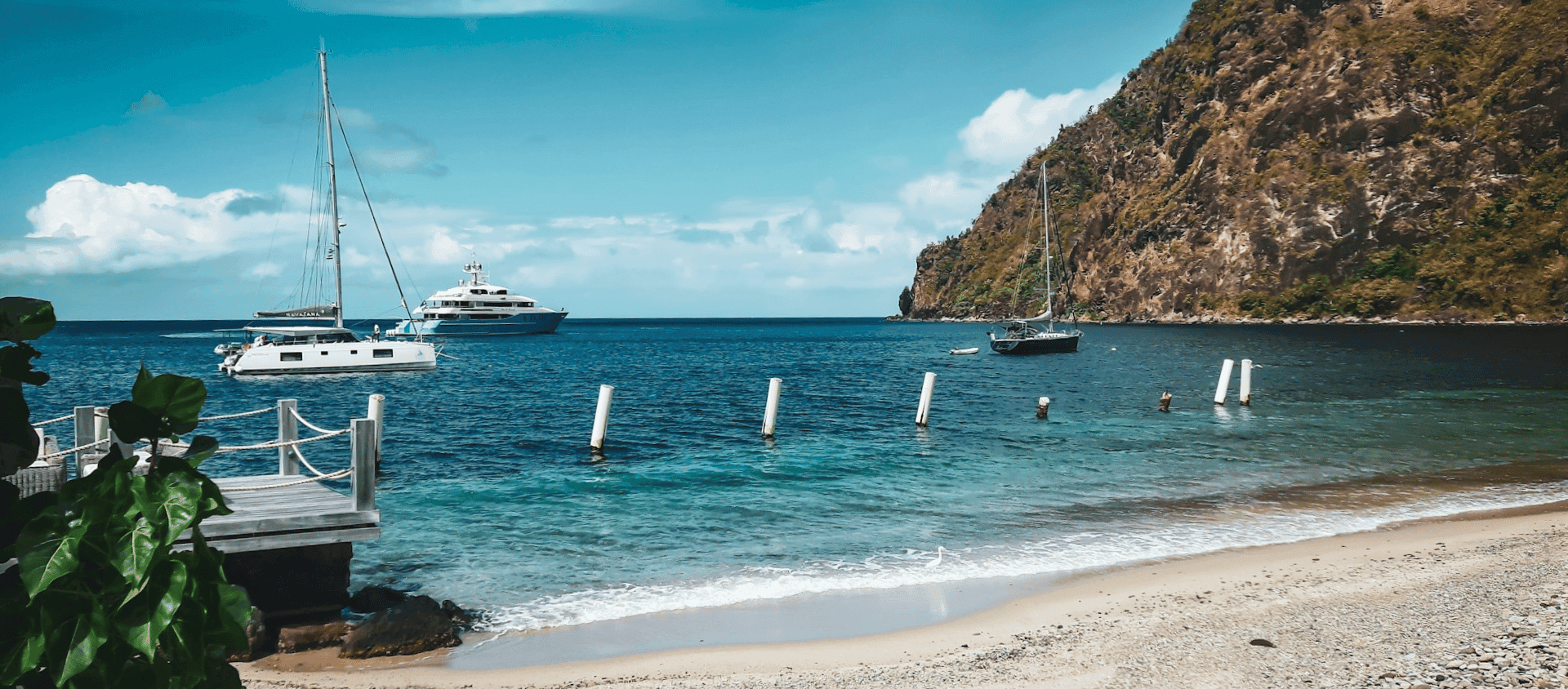 Sailboats anchored in clear blue water near a sandy beach and rocky island coastline.