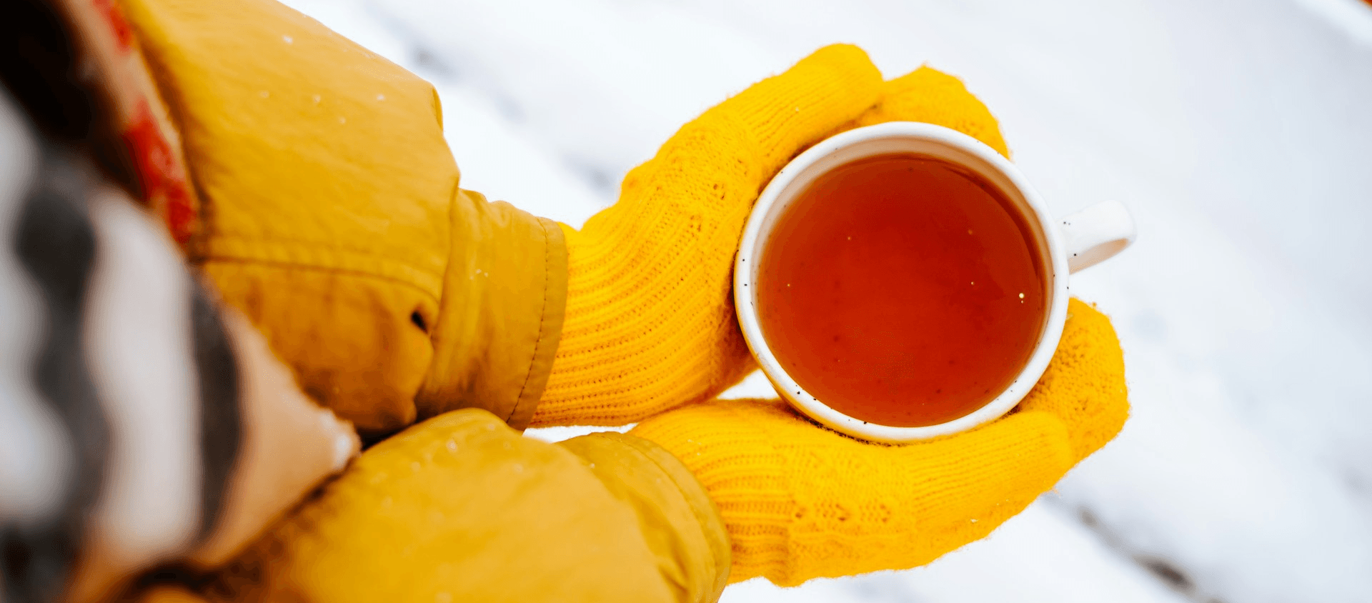 Person wearing yellow gloves holding a cup of tea outdoors in the snow.