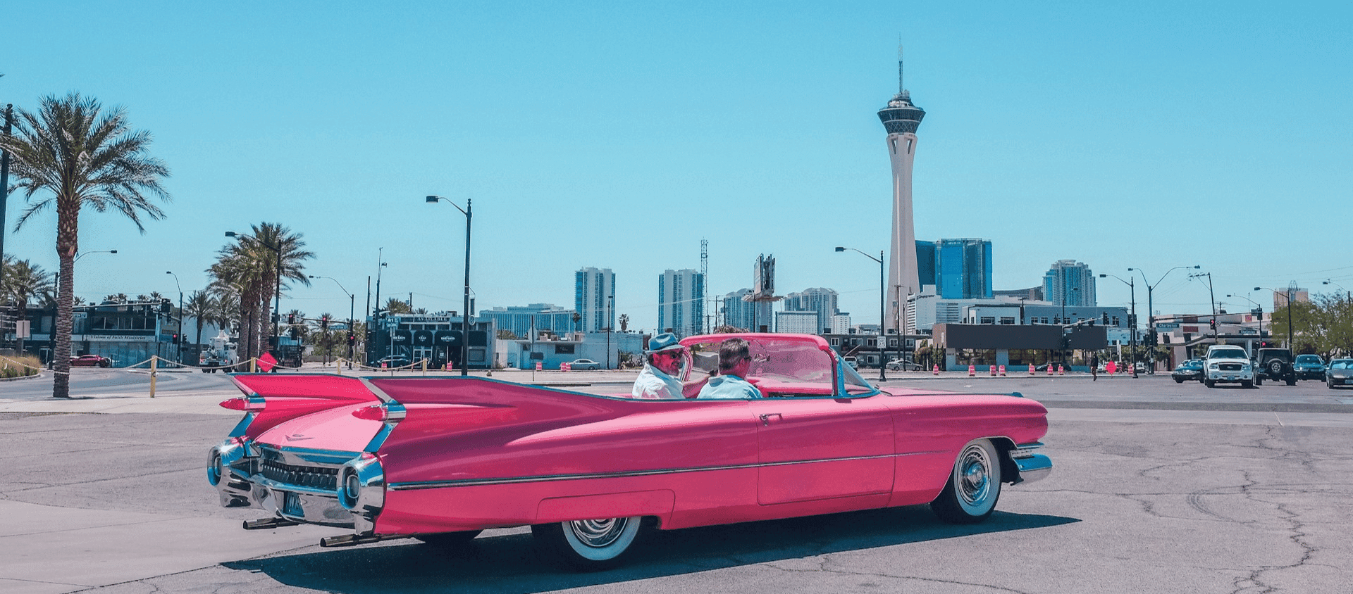 2 people driving through Las Vegas in a pink classic car