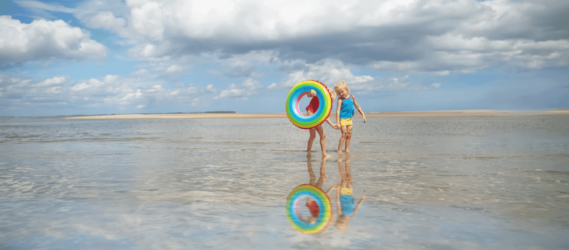 Two children walking in shallow water on a beach, one holding a colorful inflatable ring.