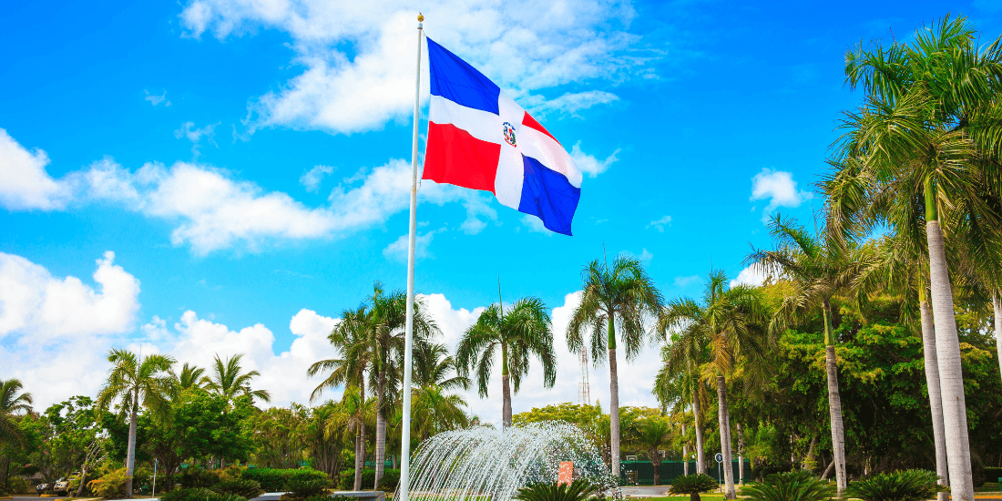 Dominican Republic flag flying on a pole above palm trees and a fountain under a blue sky.