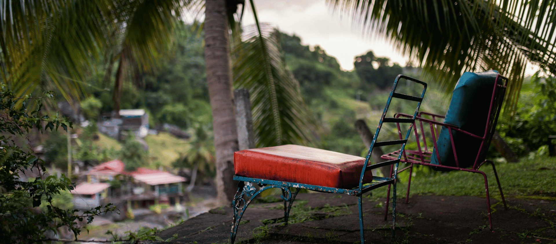 Vintage outdoor chairs beneath palm trees with a scenic view of a tropical hillside and village below.