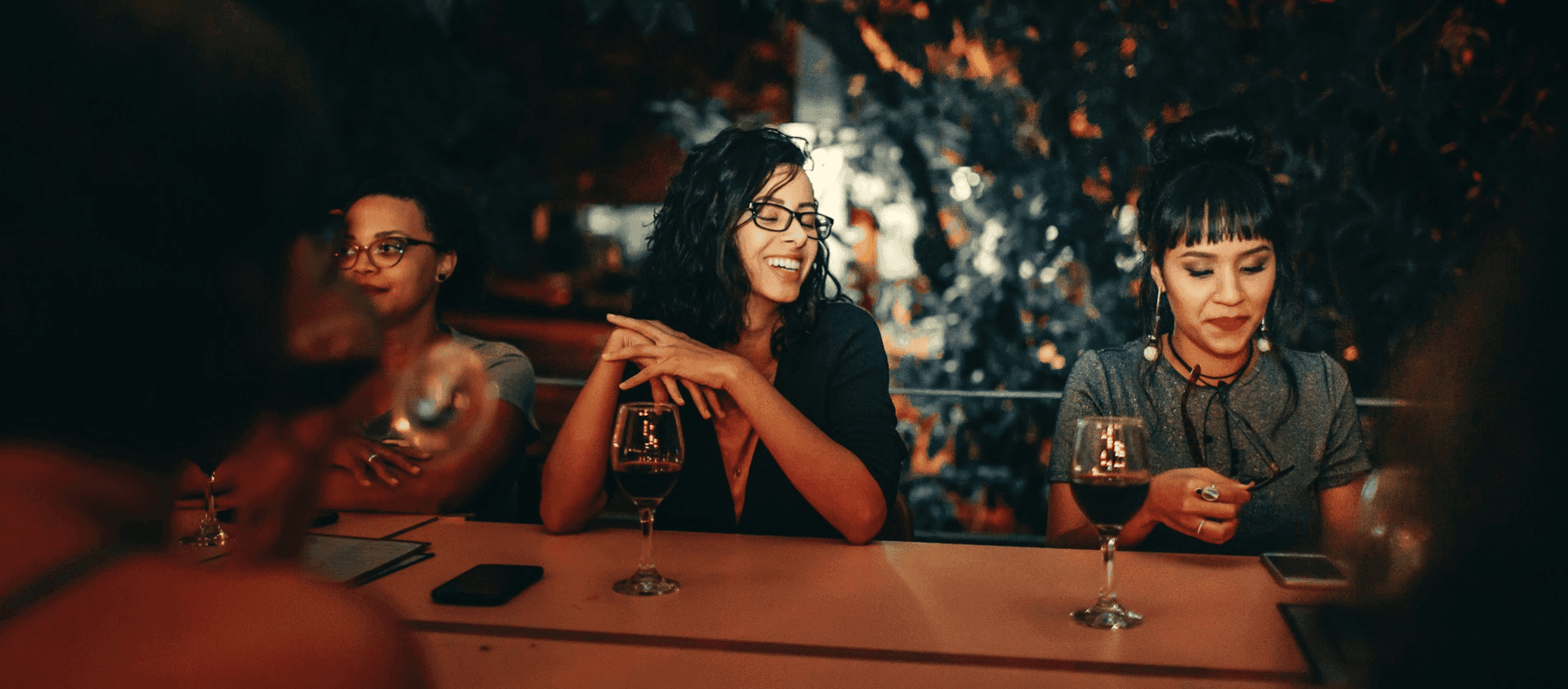 Three women sitting at a bar smiling with glasses of red wine.