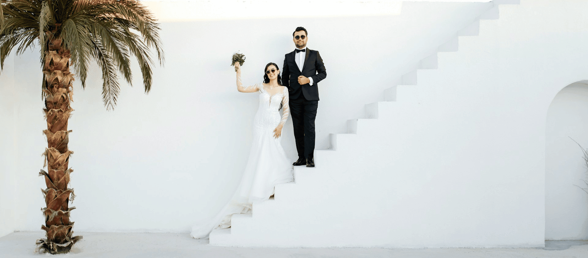 Bride and groom in formal wear posing on white steps beside a palm tree.