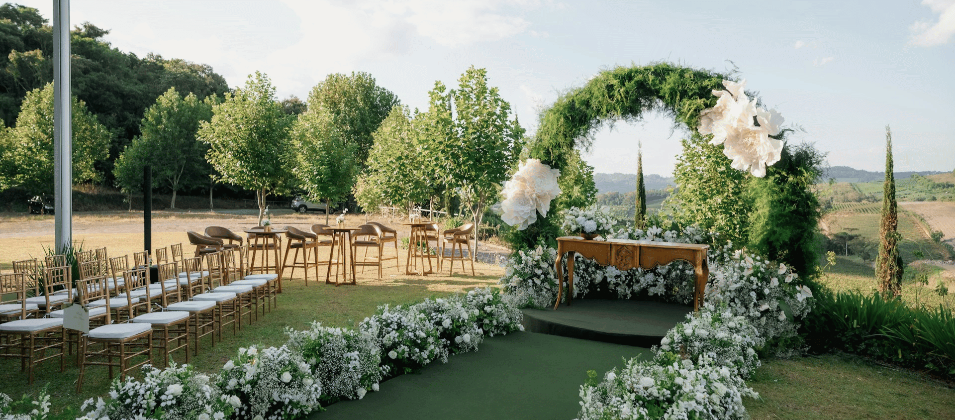 Outdoor wedding altar with greenery arch and white floral arrangements in a scenic landscape.