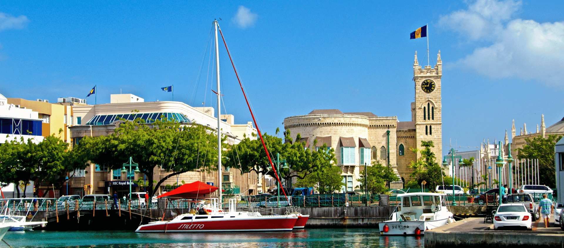 Red catamaran and yachts moored along the Bridgetown careenage in Barbados, with colorful buildings and the historic Parliament clock tower behind.