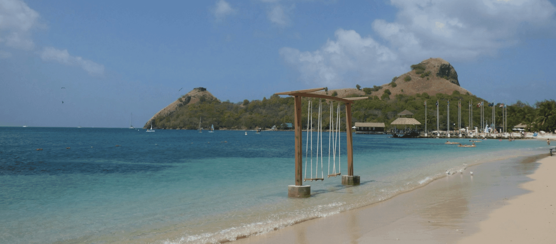 Beachfront swing over clear blue water with boats and green hills in the background.