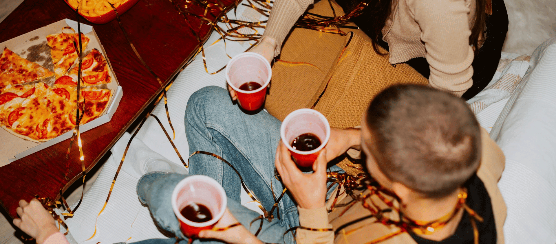 People sitting together holding red cups with juice, along with pizza and snacks on a table.
