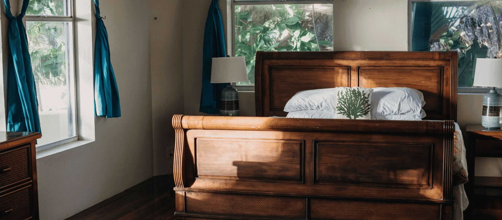 Sunlit bedroom with a wooden bed, white pillows, and blue curtains beside windows.