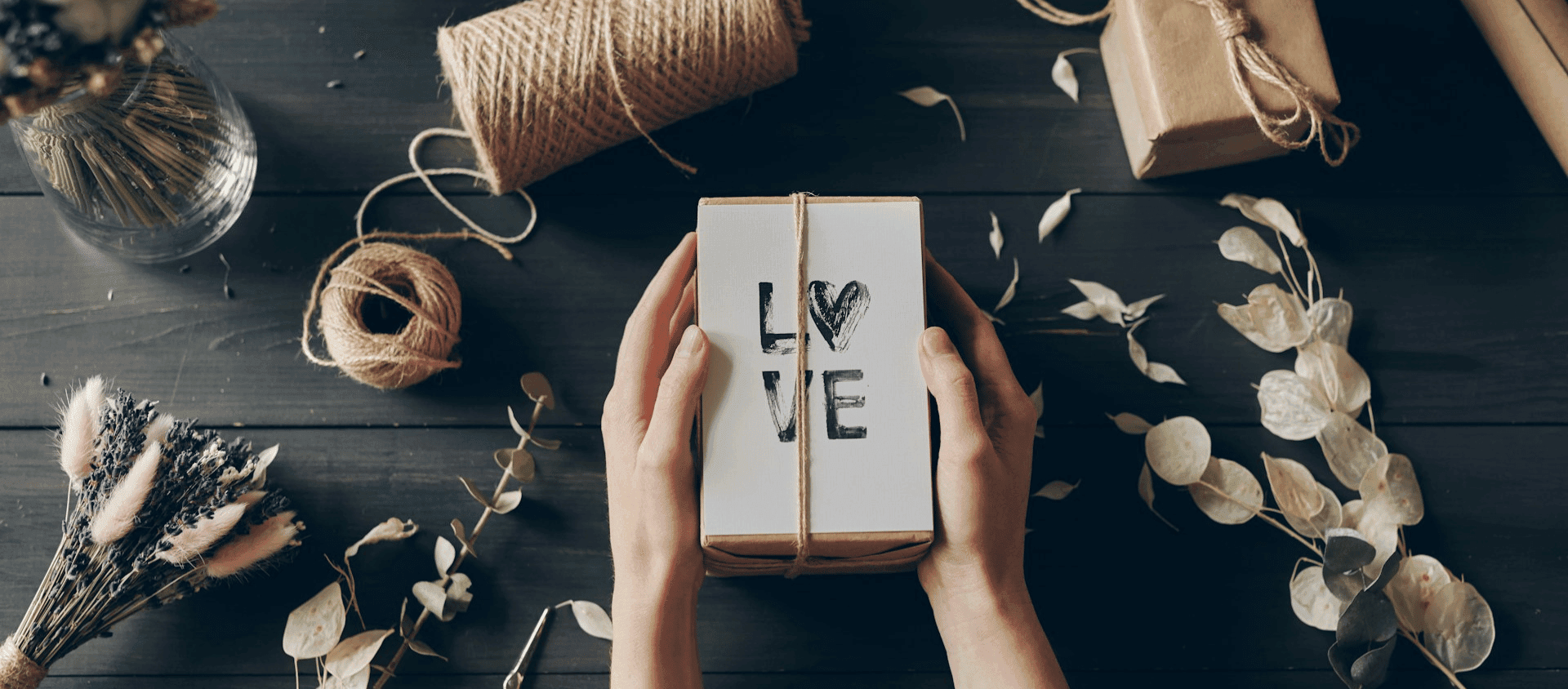 Hands holding a wrapped gift box with the word “LOVE” on the cover, surrounded by twine, flowers, and leaves on a dark wooden table.