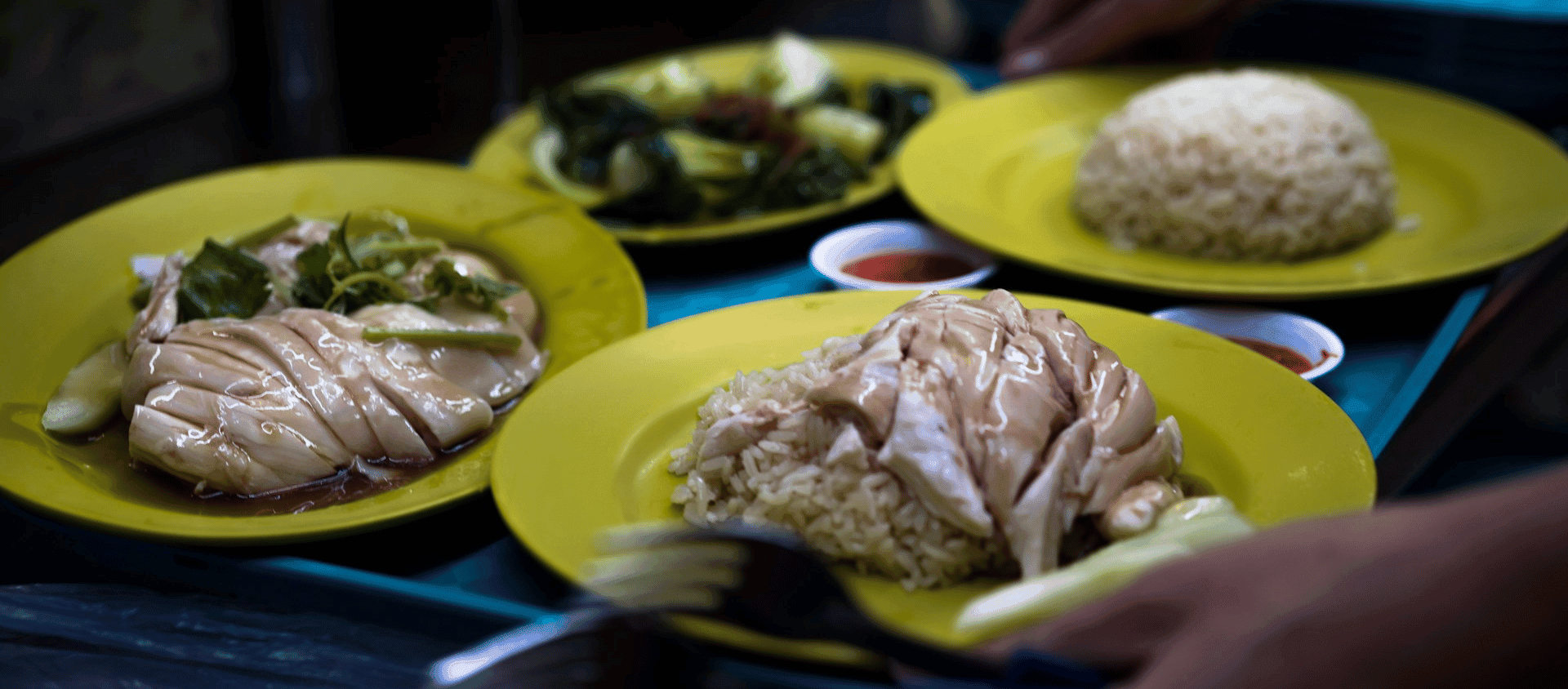Plates of Hainanese chicken rice and greens served on yellow dishes at a Singapore hawker centre