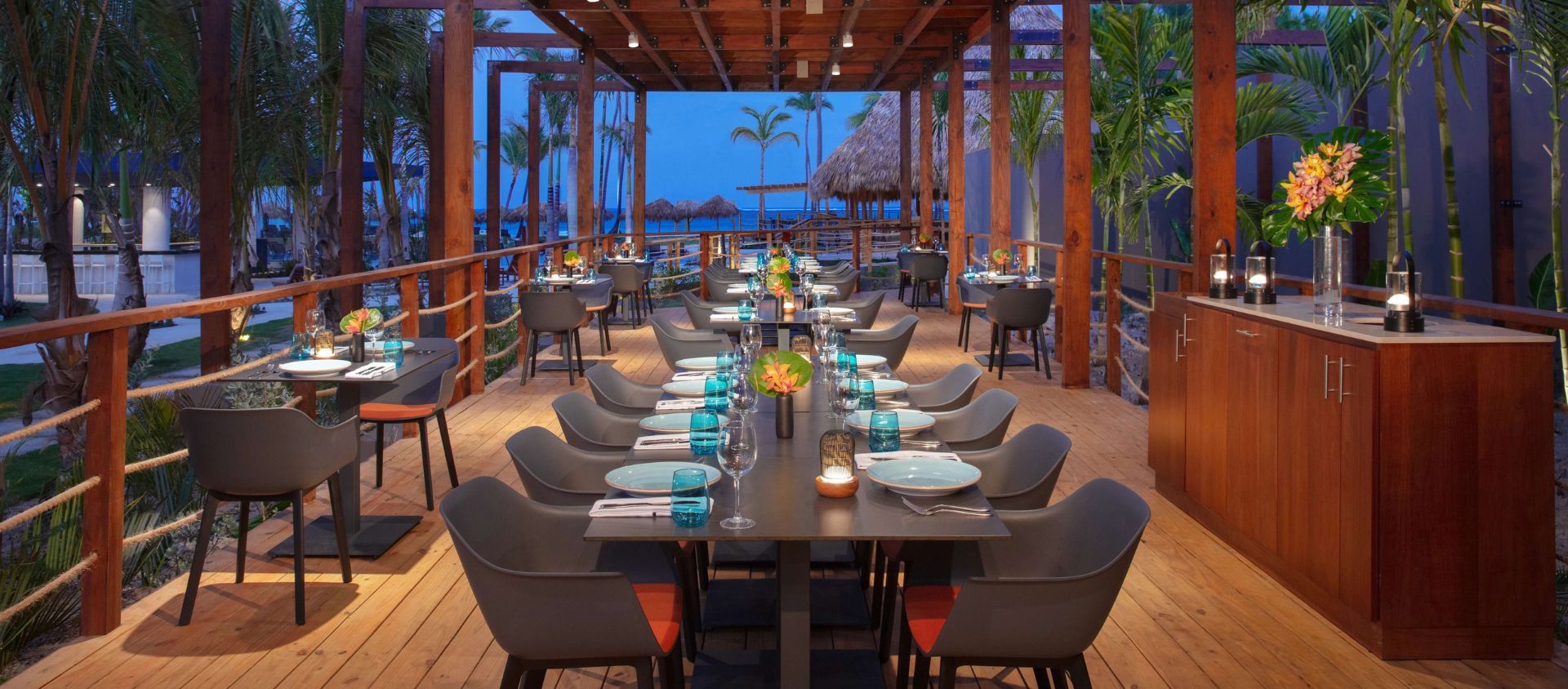 Oceanfront private dining table set for a group dinner at dusk on a wooden deck.