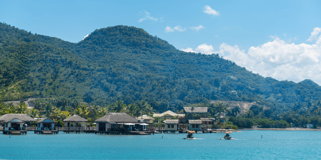 Overwater bungalows along a calm blue lagoon with green hills in the background.