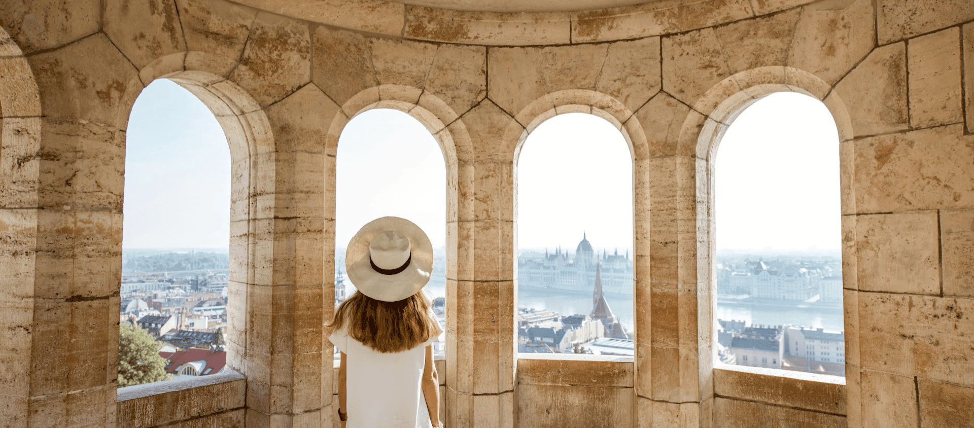 Woman in a sun hat looking through arched stone windows overlooking the Budapest skyline.