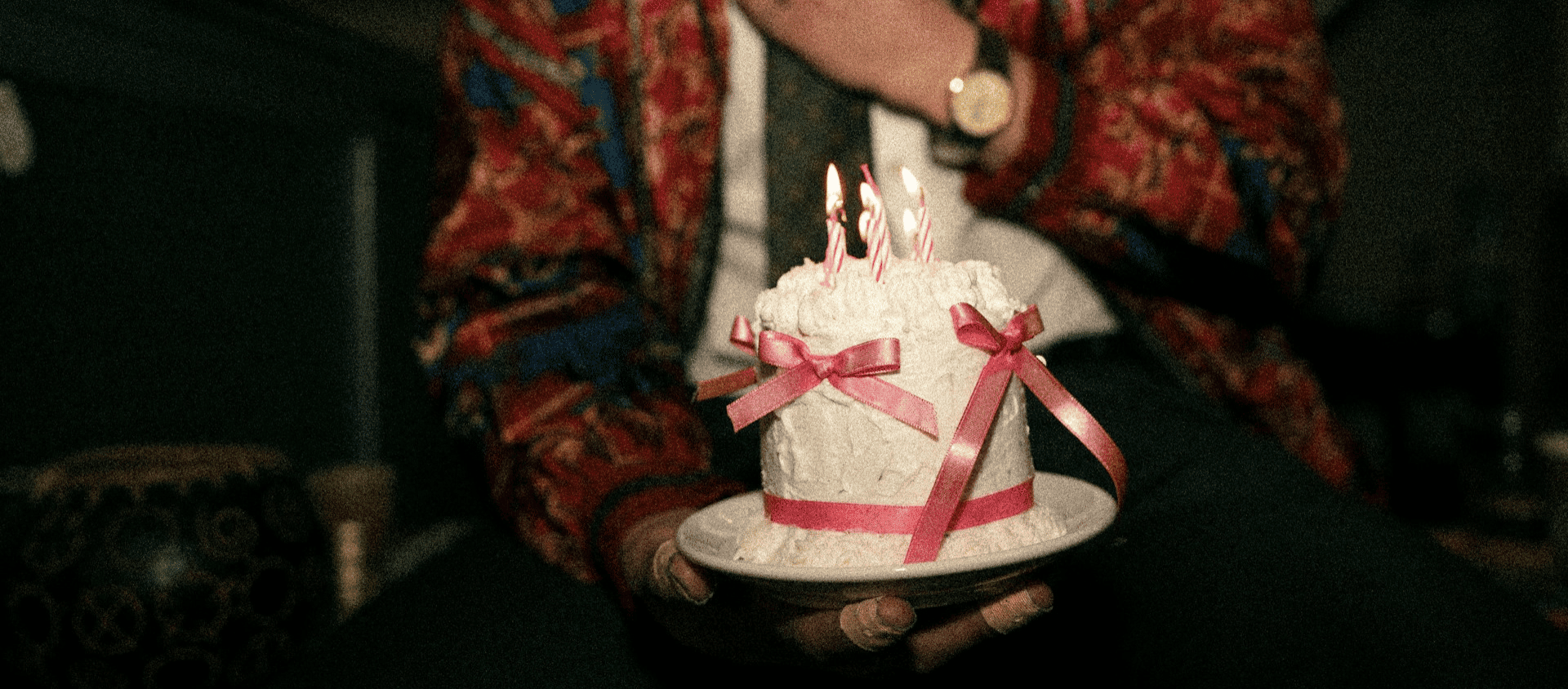 Person in a colorful outfit holding a small white birthday cake with lit candles.