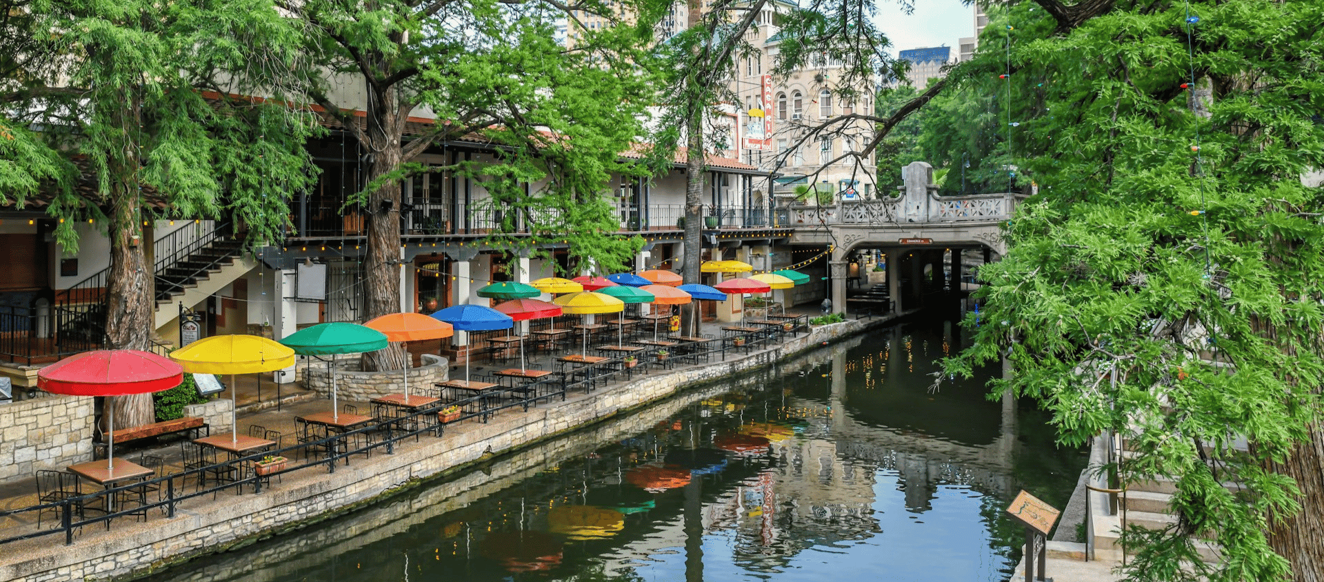 River side walkway with outdoor tables and colorful umbrellas beside a canal.