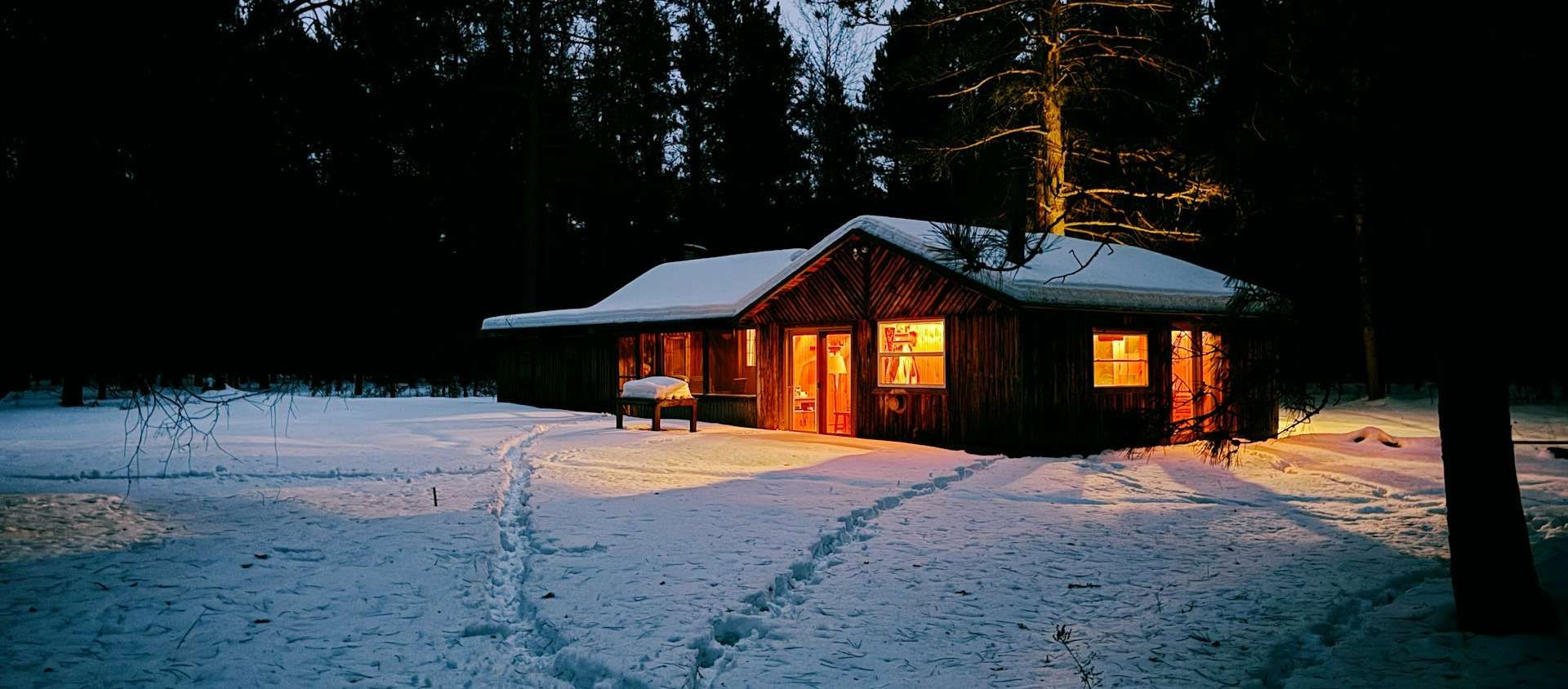 A cozy wood cabin glowing with warm light at dusk, surrounded by snowy pines with footprints leading to the door.
