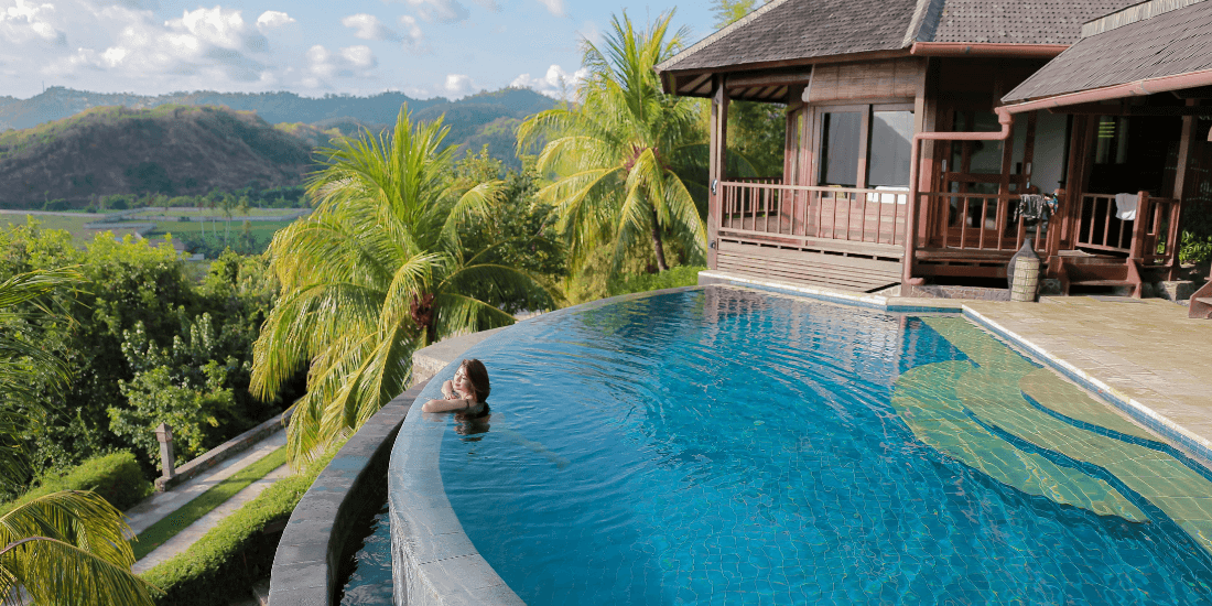 Person relaxing in an infinity pool overlooking palm trees and green hills.