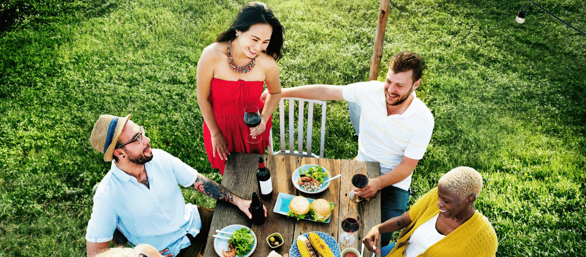 Group of friends sitting outdoors sharing food and drinks on grass.