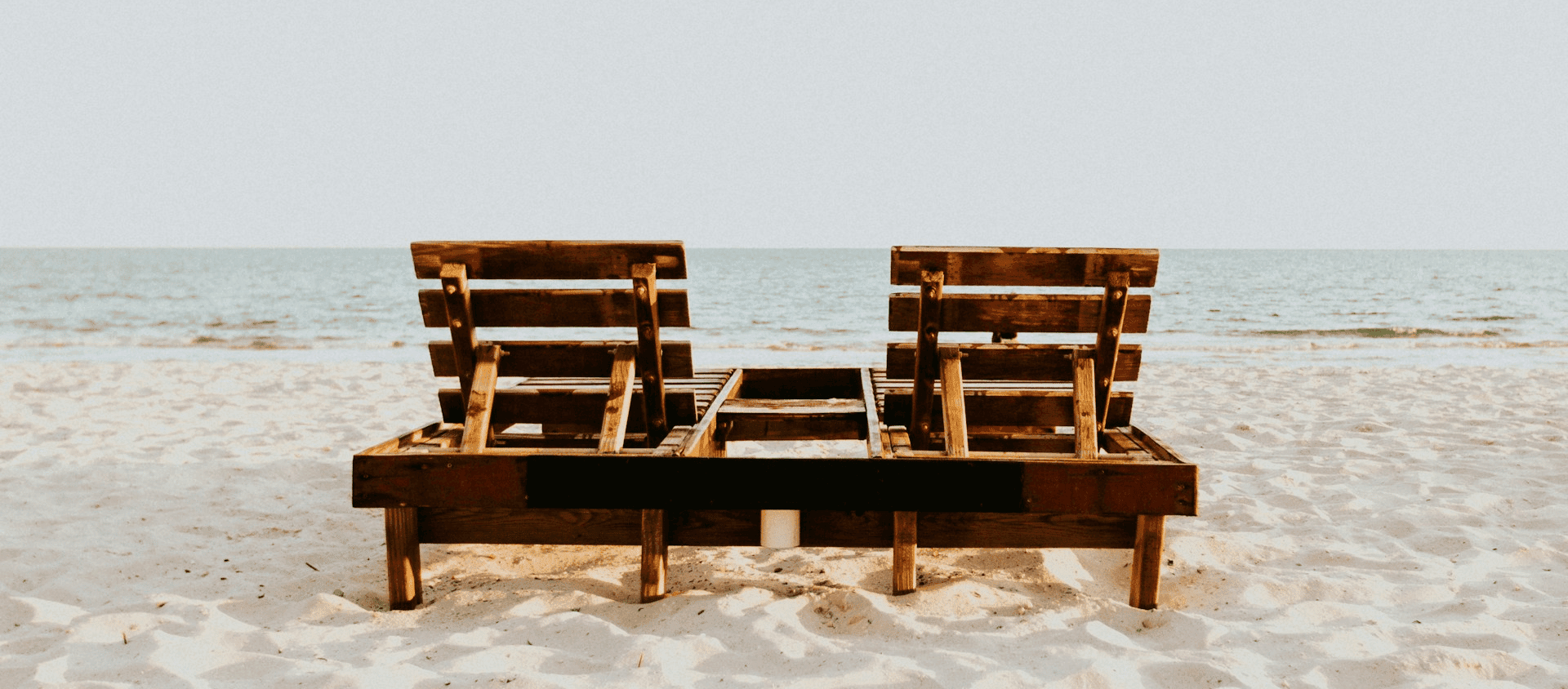 Two wooden beach lounge chairs sitting on sand facing the ocean.