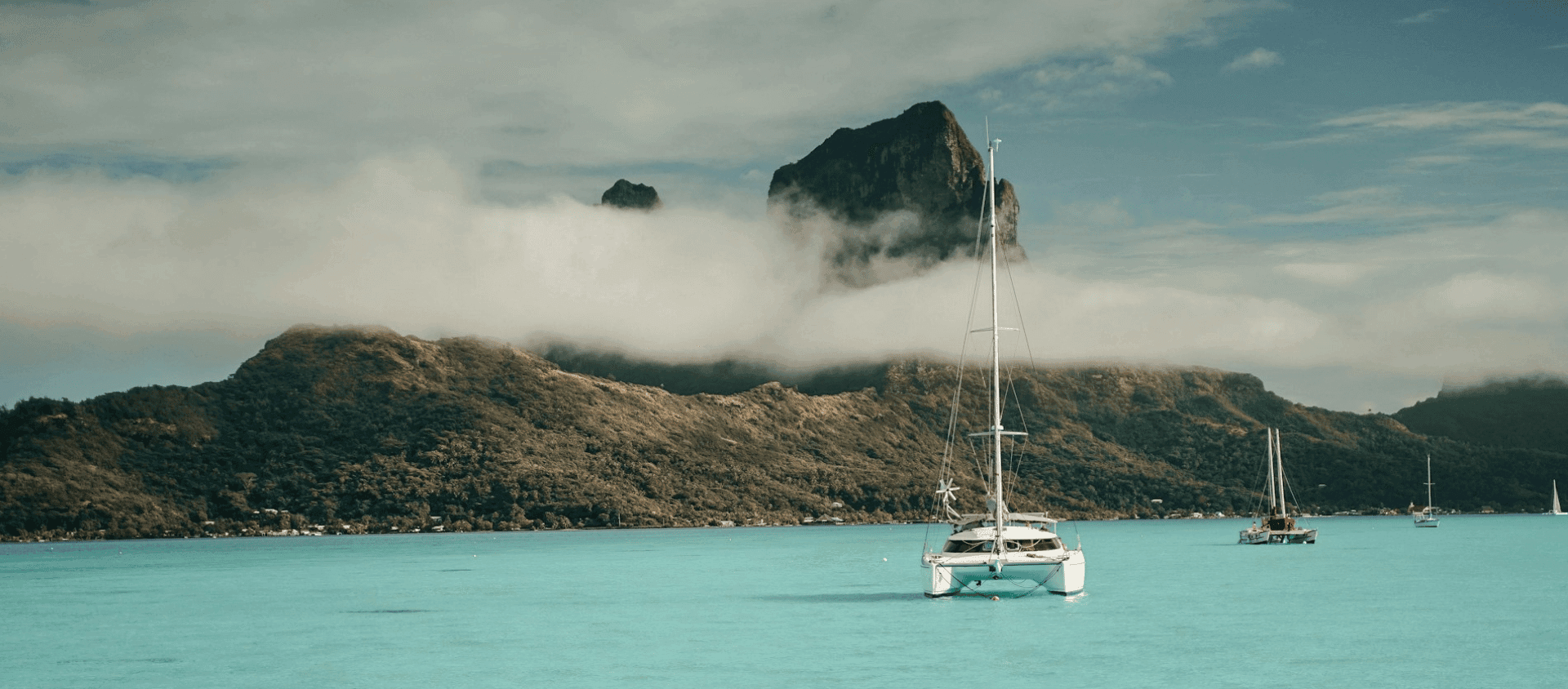 Sailboats anchored in a turquoise lagoon with a mountain rising above low clouds in Bora Bora.