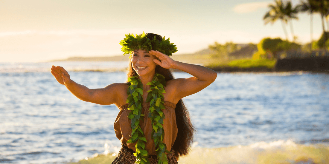 Hula dancer wearing a leaf lei performs on a beach at sunset.