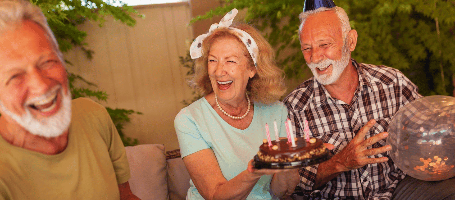 Three elderly people laughing while celebrating with a birthday cake outdoors.