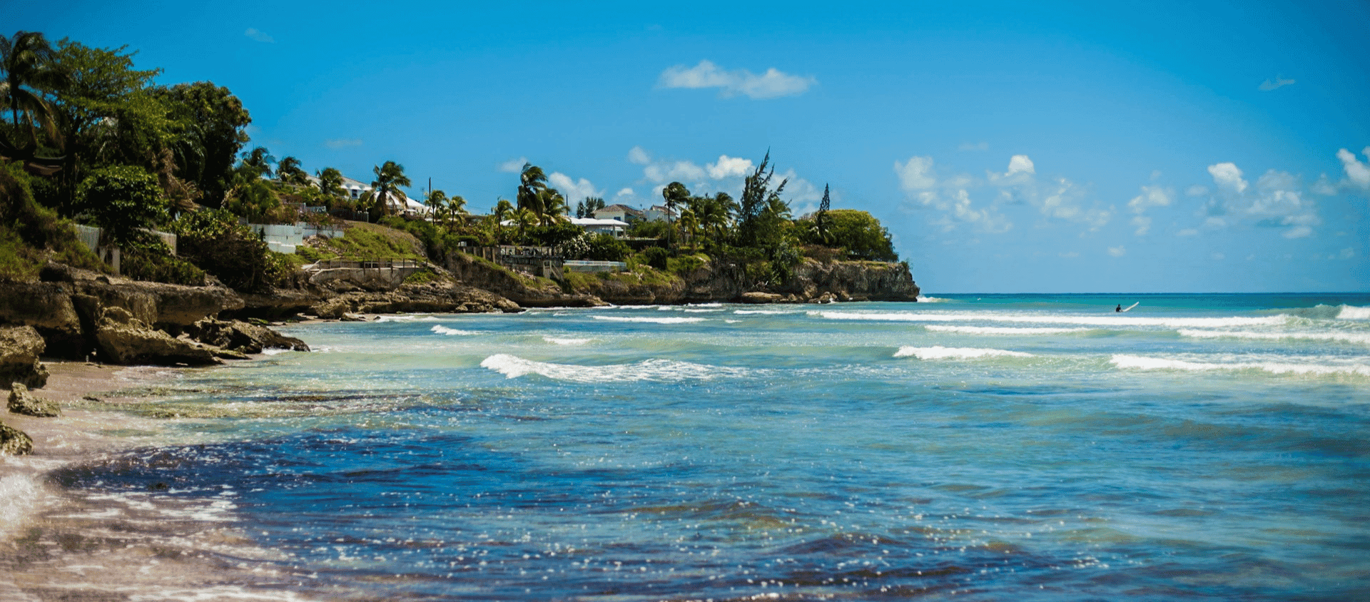 Rugged Barbados coastline with Atlantic surf rolling onto a sandy beach and cliffside villas tucked into green hills.