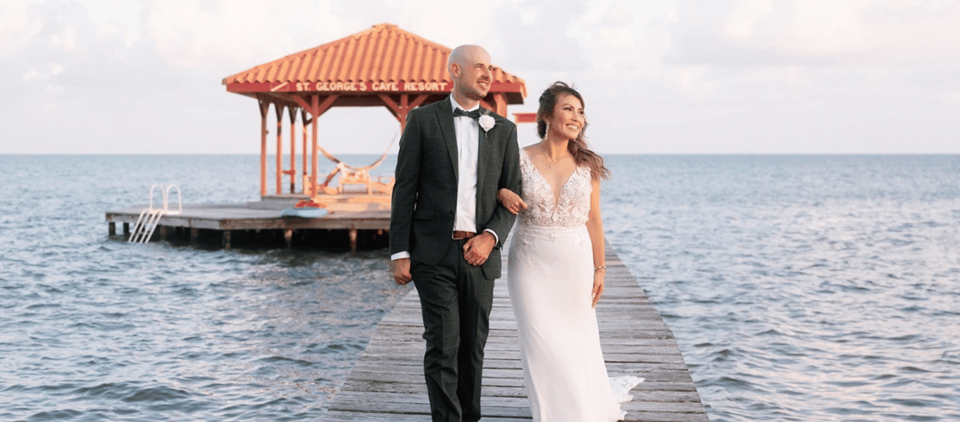 Newlywed couple walking along a wooden dock surrounded by calm water.