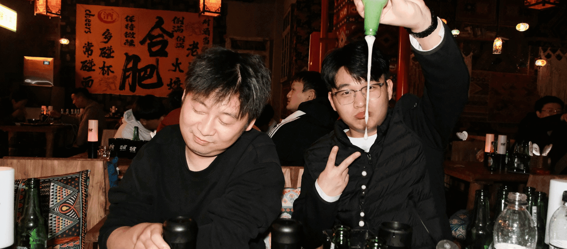 Two men at a restaurant, one playfully pouring a beverage.