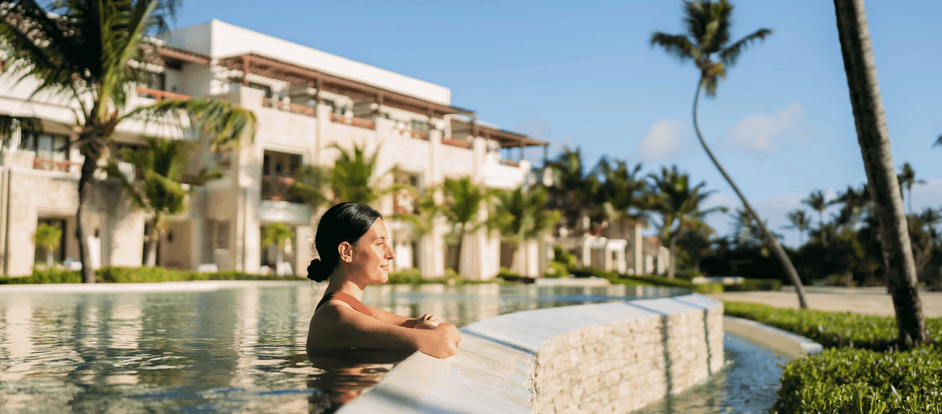 Woman relaxing in a resort pool with luxury suites and palm trees in the background at Secrets Cap Cana.