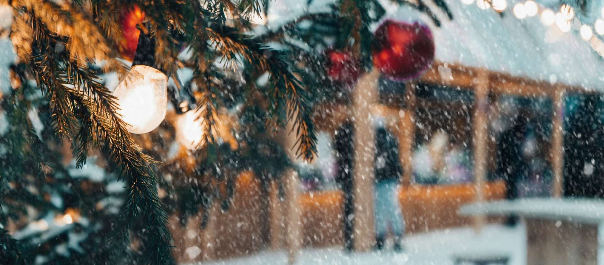 Snow falling on evergreen branches strung with warm string lights and red ornaments, with a lit patio blurred in the background.
