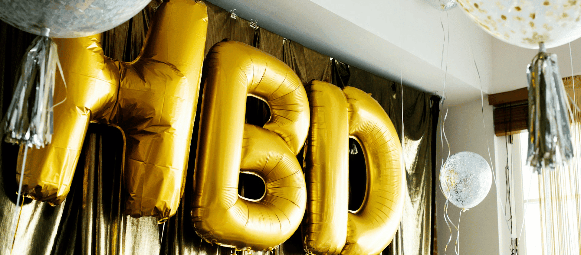Gold balloon letters spelling “HBD” with party decorations in a room.